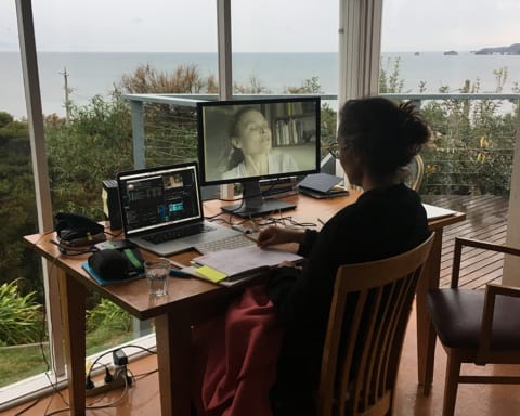A woman sits with her back to the camera at a desk with a computer editing station in a room with floor to ceiling windows, beyond which lies a green forest and the water horizon of Waratah Bay in Gippsland, Australia.